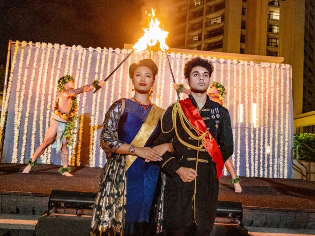 The young man and woman representing Hawaiian royalty on stage as performers light up torches behind them at Nā Lei Aloha lūʻau.