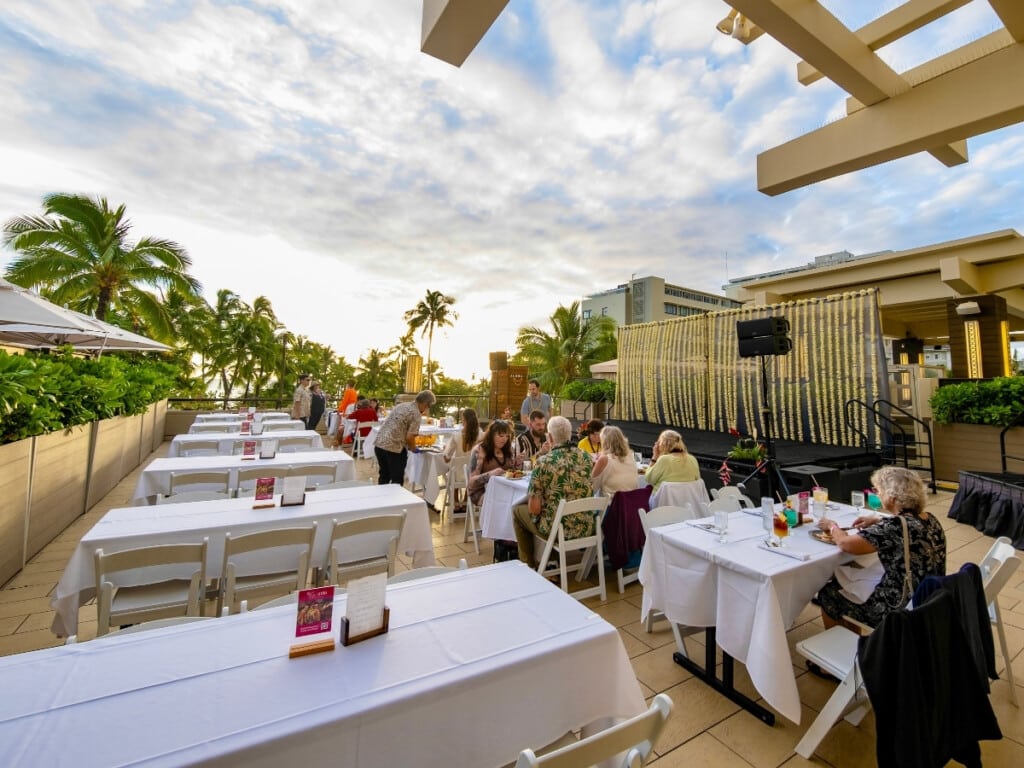 The intimate open air setting at the Nā Lei Aloha lūʻau which includes tables lined up in front of a stage with the palm trees and the sunset in the background.