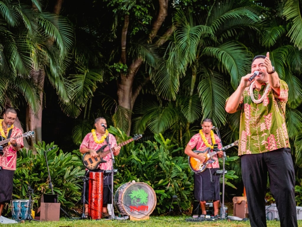 Pre-show performance at Waikiki Starlight Luau: Ocean Lawn, featuring a Hawaiian band in colorful aloha shirts and lei playing live music surrounded by tropical greenery.
