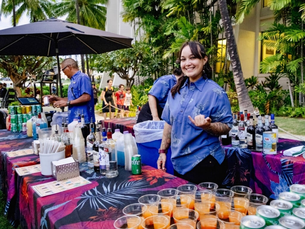 Bar setup with mai tais t Waikiki Starlight: Ocean Lawn with the bar tender wearing blue aloha shirt. She is smiling and making a shaka sign.