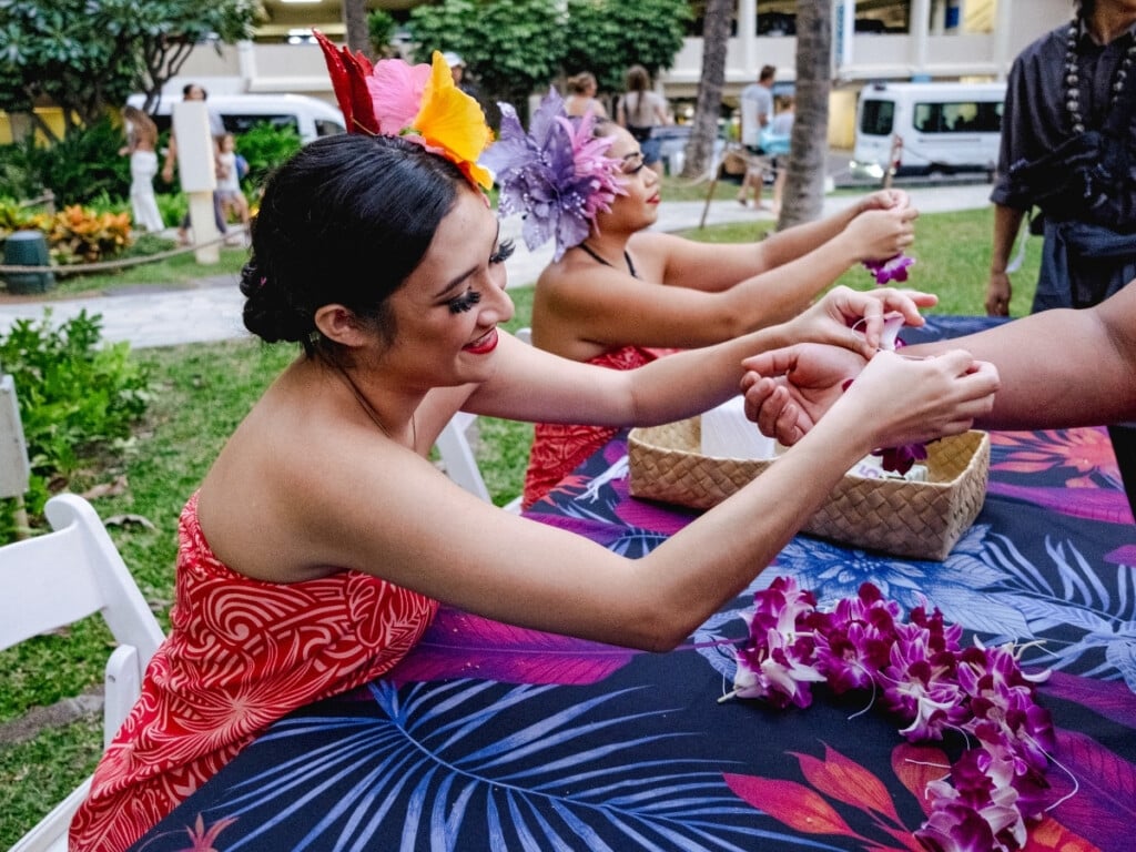 Two ladies making bracelet leis made with fresh purple orchids for guests at Waikiki Starlight Luau: Ocean Lawn.