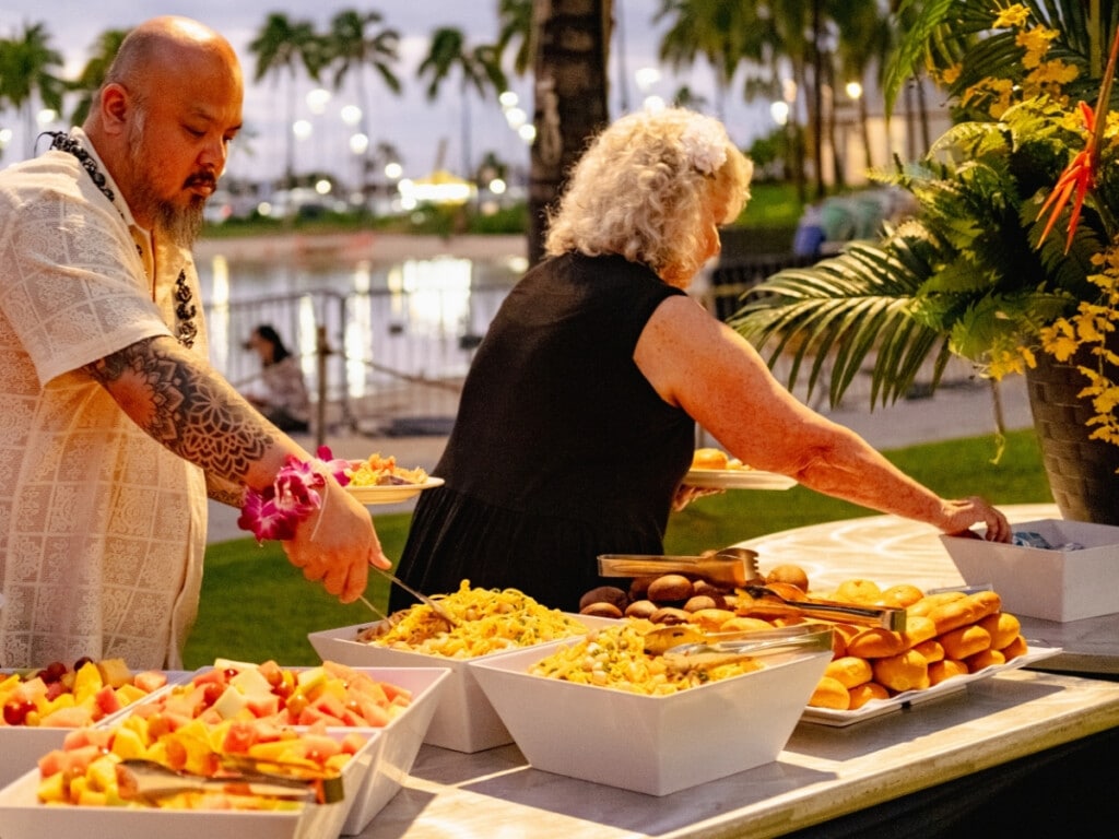 Guest serving themselves at the buffet of Waikiki Starlight Luau: Ocean Lawn with sunset and tropical scenery in the background.