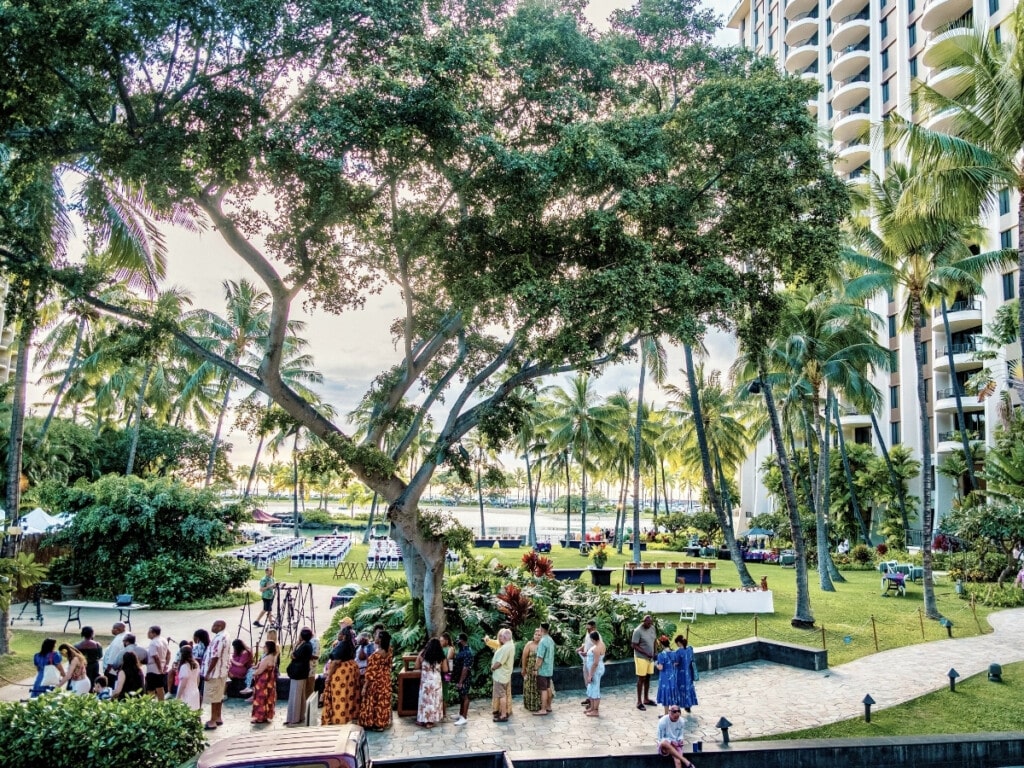 Outdoor view of the Starlight Luau Ocean Lawn at the Hilton Hawaiian Village, featuring lush palm trees, a large canopy of greenery, and neatly arranged tables on the lawn. Guests dressed in colorful Hawaiian attire gather along a paved walkway, surrounded by tropical landscaping and overlooking the ocean.