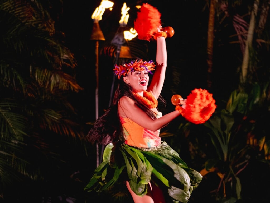 Hawaiian hula dancer performing at the Waikiki Starlight Luau: Ocean Lawn adorned with a colorful flower crown and traditional attire set against a tropical background with tiki torches.