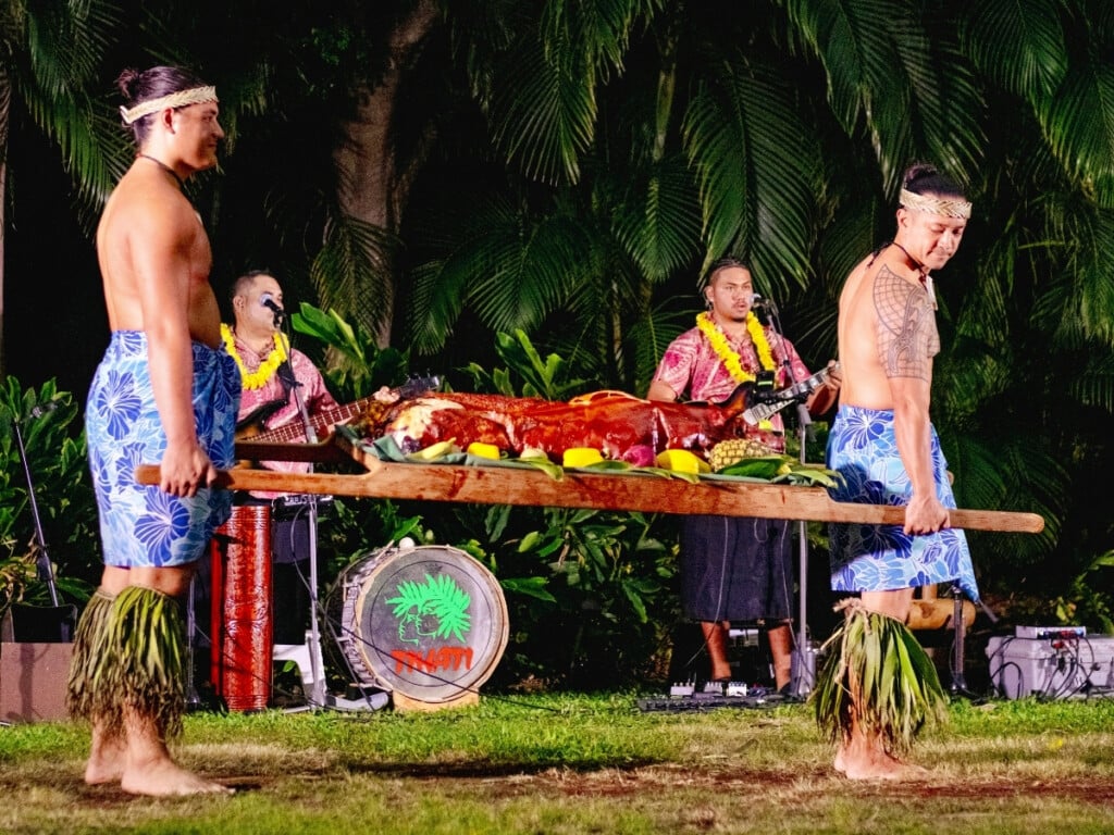 Performers at Starlight Luau carrying a roasted pig on a large wooden platter as the Hawaiian band plays in the background with lush tropical greenery surrounding the scene.