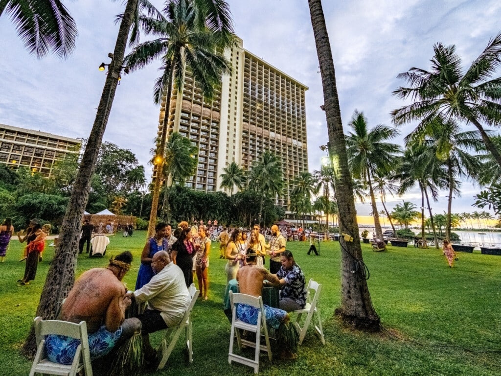 Luau guests getting temporary Polynesian tattoos - a pre-luau activity on the lawn at Hilton Hawaiian Village at the Waikiki Starlight Luau: Ocean Lawn featuring palm trees with the sun setting over Duke Kahanamoku Lagoon in the background.