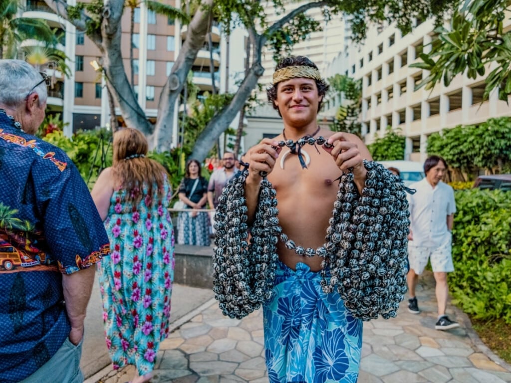 A lei greeting at the Waikiki Starlight Luau Ocean Lawn at Hilton Hawaiian Village in Waikīkī, O‘ahu with a man in traditional Polynesian attire holding a black and white kukui nut lei.