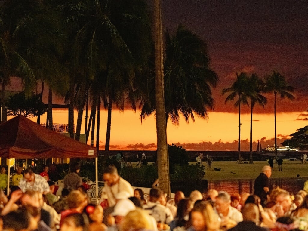 View of vibrant sunset near Duke Kahanamoku Lagoon with the guests seated in the foreground at the Waikiki Starlight Luau: Ocean Lawn.
