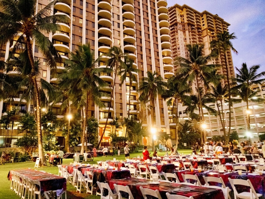 Colorful table set up on the lawn at Waikiki Starlight Luau: Ocean Lawn Photos in Hilton Hawaiian Village featuring palm trees and the hotel in the background.