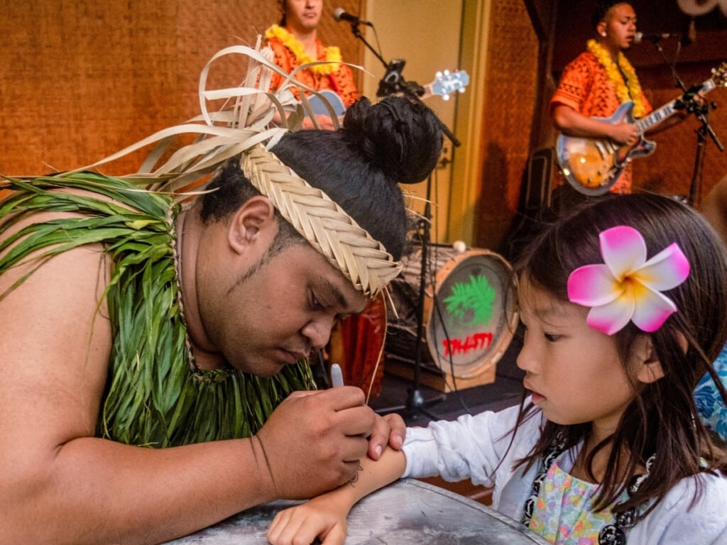 A little girl with a flower in her hair getting a temporary Polynesian tribal tattoo with the band in the background at the Waikiki Starlight Luau: Rooftop.