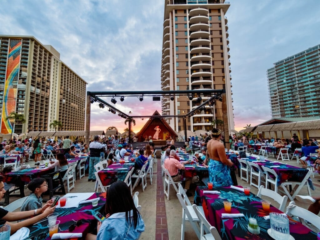 Guests watching the MC and the band perform at Waikiki Starlight Luau: Rooftop with Rainbow tower in the background.