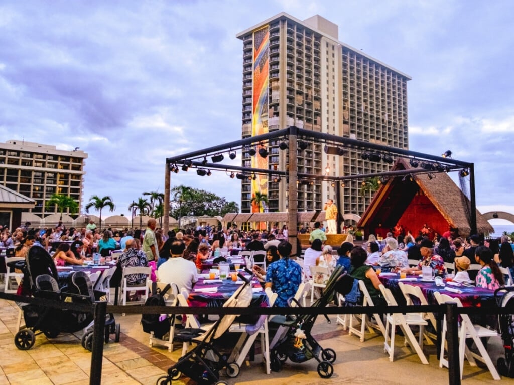 Guests watching the MC and the band perform at Waikiki Starlight Luau: Rooftop with Rainbow tower in the background.