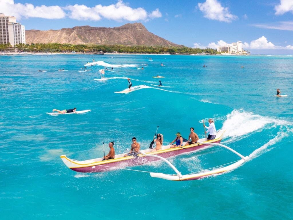 A Hawaiian outrigger canoe riding a wave towards Waikīkī Beach.