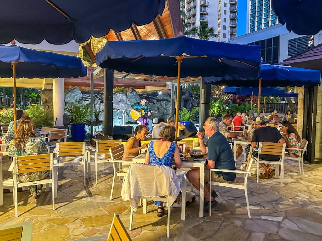 Live music performance at Kani Ka Pila Grille in Outrigger Reef Waikiki Beach Resort, featuring a guitarist entertaining diners in an inviting outdoor seating area under navy blue umbrellas.