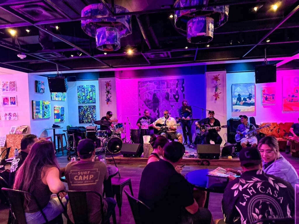 A six-member band performing at The Secret Spot Honolulu, with three guitarists, a keyboard player, and two drummers, all illuminated by vibrant fuchsia lighting as the audience watches the lively show.