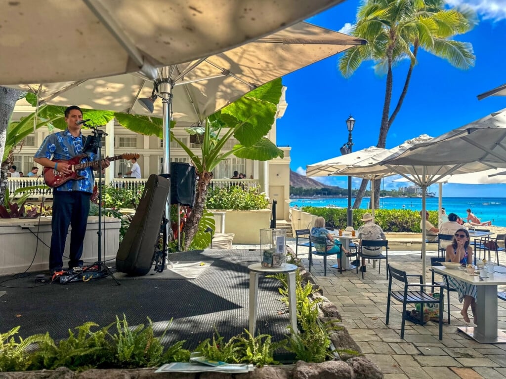 Musician performing with a guitar and singing at The Beach Bar of Moana Surfrider, a beachfront venue on Waikīkī Beach, with a backdrop of clear blue sky, ocean waves, swaying palm trees, and a glimpse of Diamond Head in the distance.