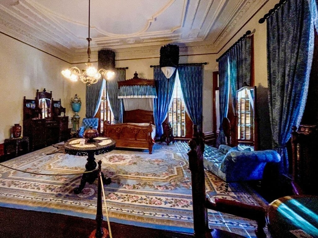 King Kalākaua’s bedroom with a mixture of blue American, Hawaiian, Asian and European furnishings. The room has floor to ceiling windows and blue curtains.
