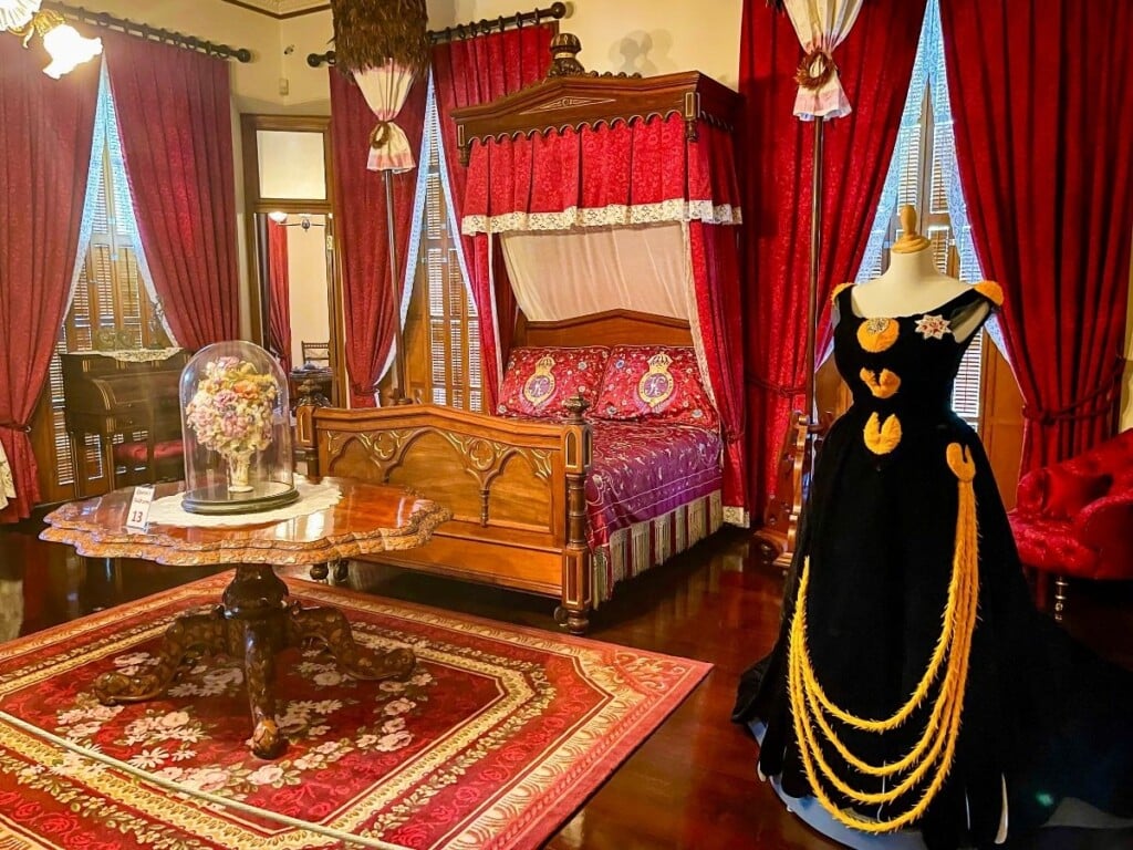 Queen Kapiʻolani’s bedroom in ʻIolani Palace which is furnished with mahogany furniture and red curtains and upholstery. It displays one of her navy blue dresses.