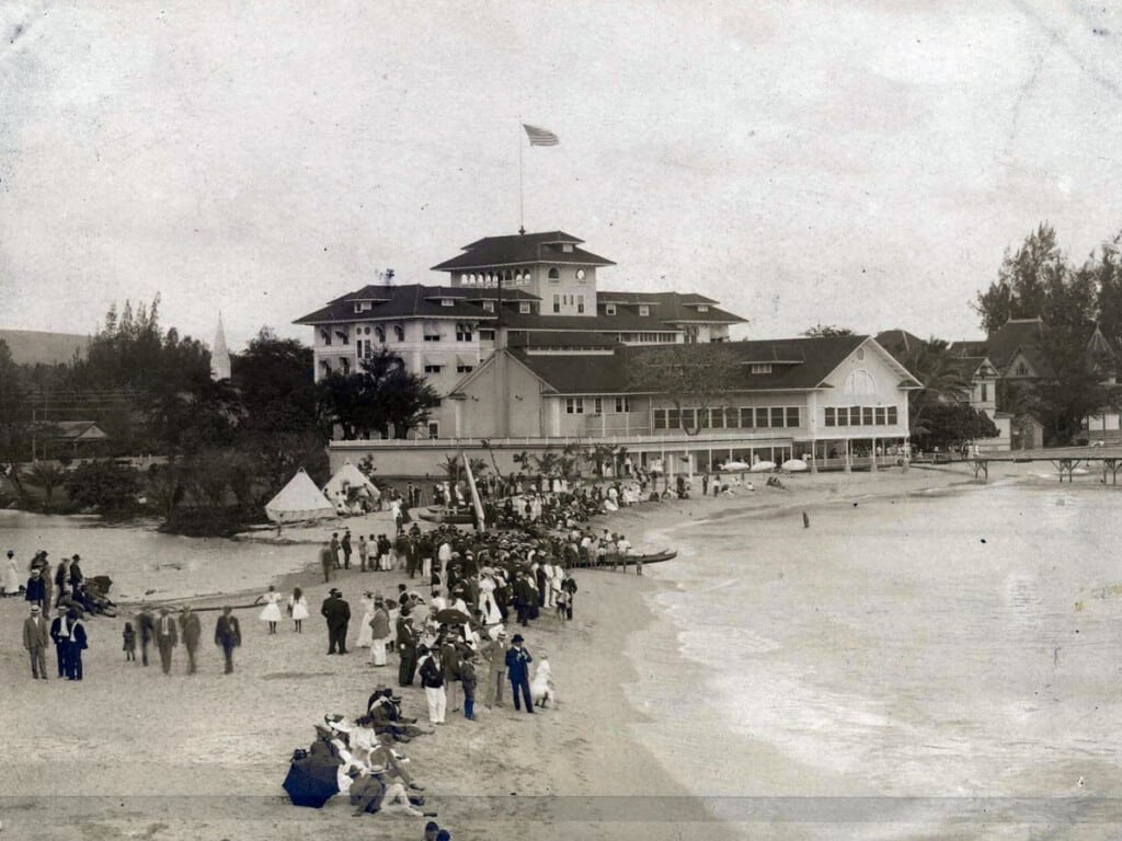 Outrigger Canoe Club members in suits and ties watch canoe races near the mouth of the ʻApuakēhau Stream. Moana Hotel is in the background. 