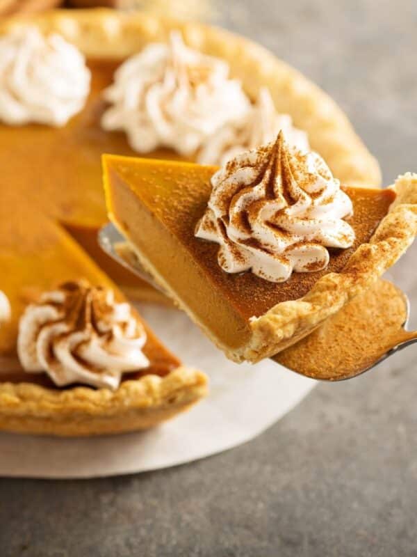 Slice of pumpkin pie topped with whipped cream and cinnamon, served from a whole pumpkin pie for Thanksgiving.