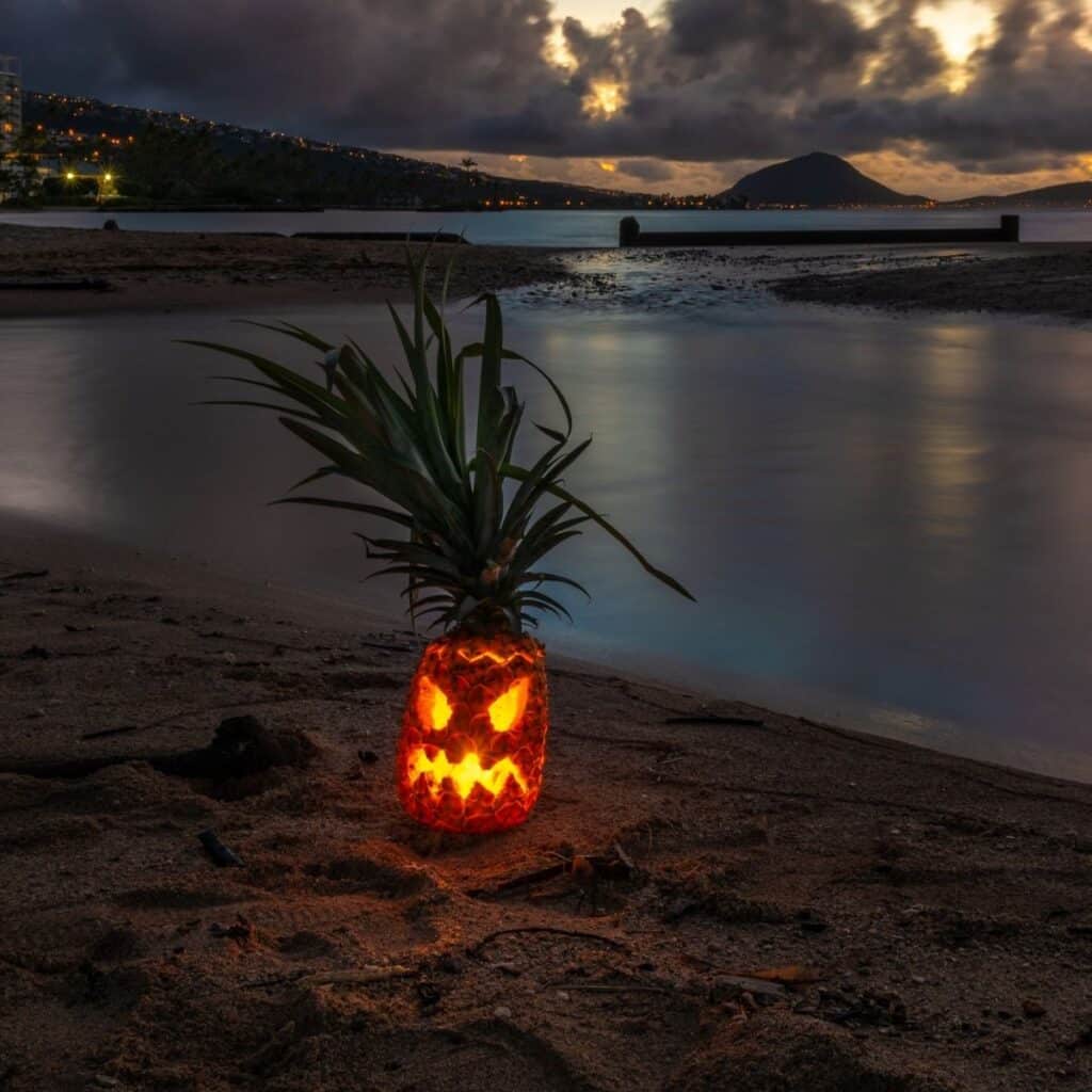Glowing jack-o’-lantern carved from a pineapple on an Oʻahu beach at dusk, with Koko Head in the distance.