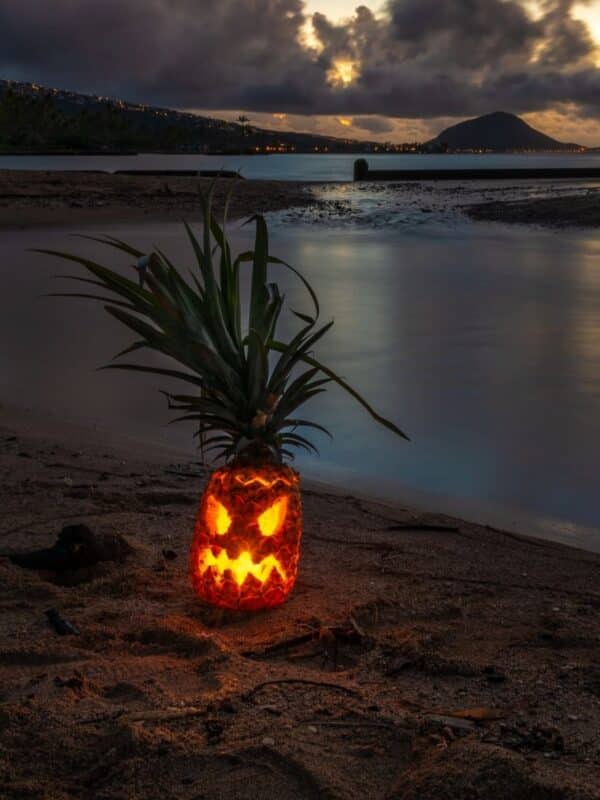 Glowing jack-o’-lantern carved from a pineapple on an Oʻahu beach at dusk, with Koko Head in the distance.
